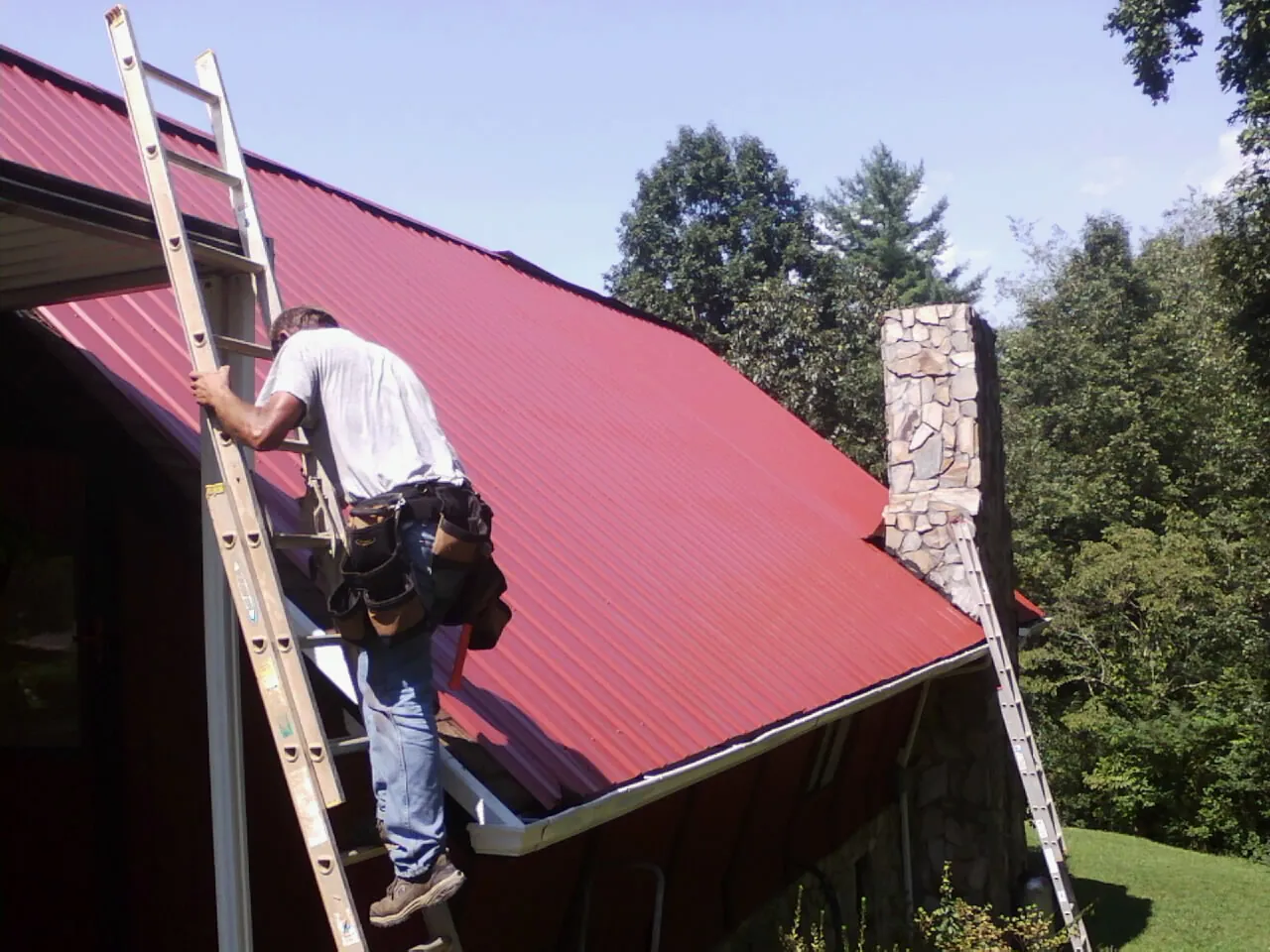 Roofer on a ladder beside a red metal roof and stone chimney — Soffit & Fascia Repair by Roofing Brothers in Grasonville